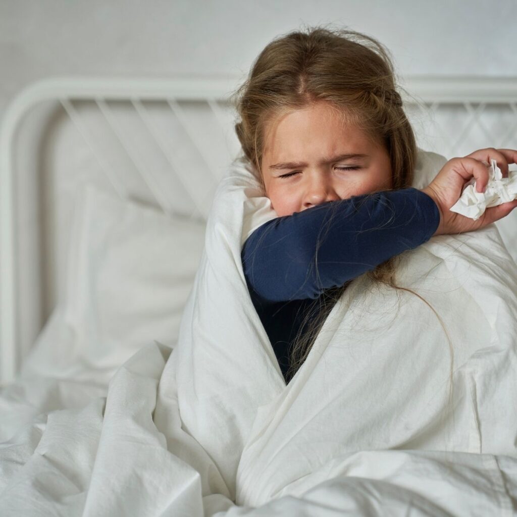 young girl sitting in bed coughing and sneezing with a tissue in her hand