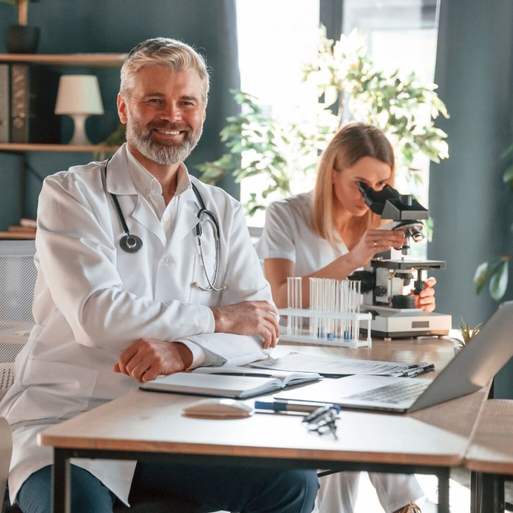 two doctors sitting at a desk. Male doctor smiling. Female doctor looking into microscope