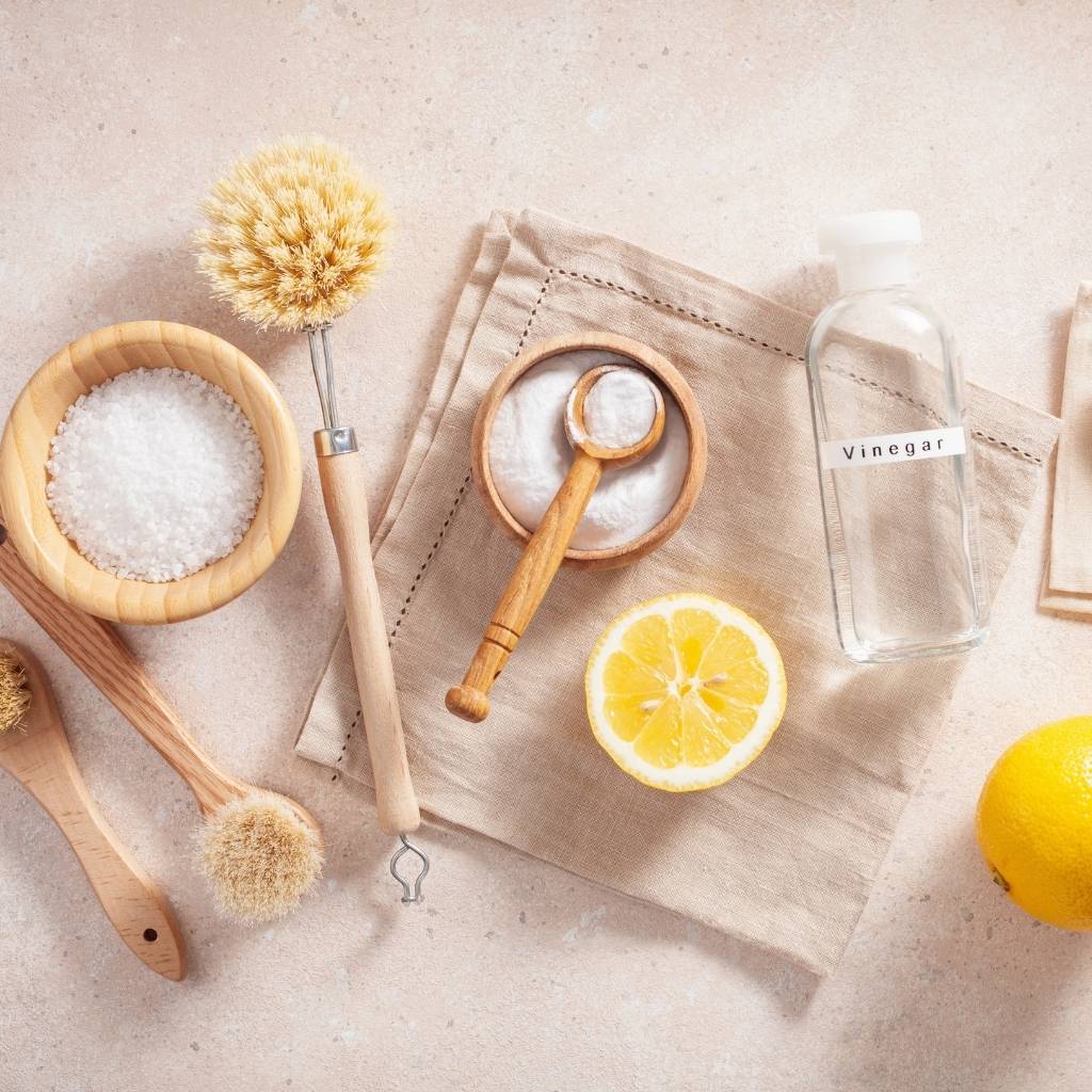 several cleaning items laying on a table including lemons, brushes, bowl of baking soda and bottle of vinegar