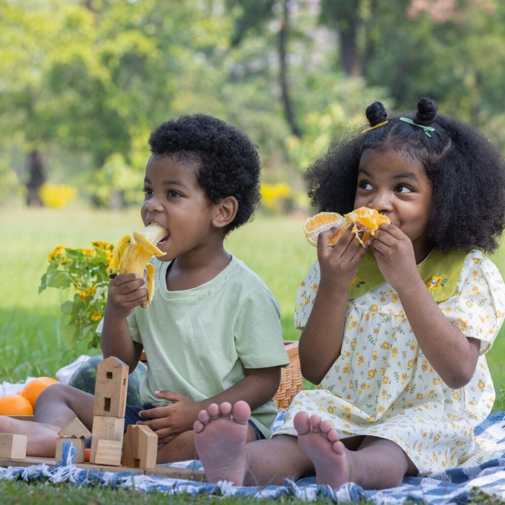 two children sitting outside on a blanket enjoying a healthy snack