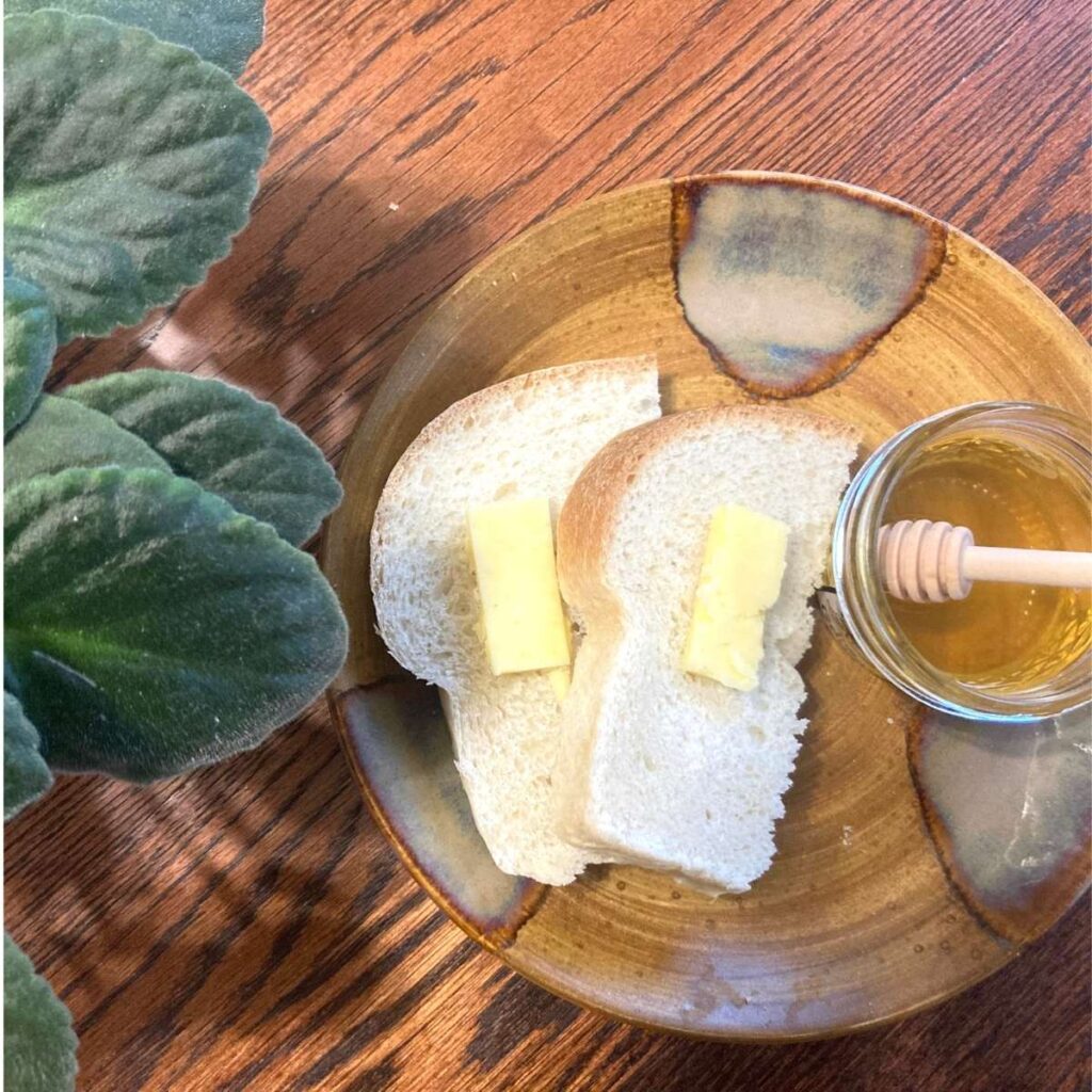 Plate with sourdough bread on it topped with butter and a small glass jar of honey with a honey spoon in it to the side. 