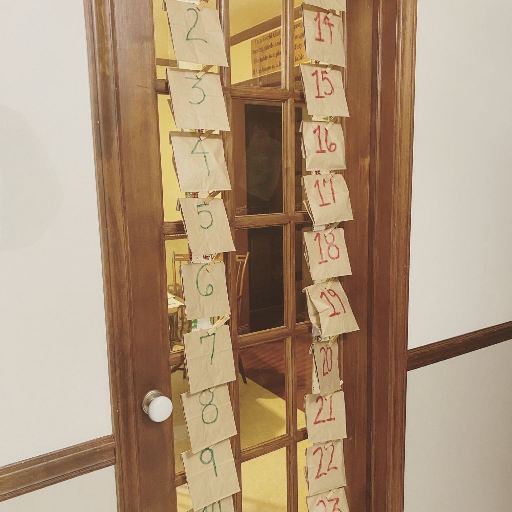 twenty four brown paper bags hanging on a french door with numbers one through twenty four written on them to be an advent calendar